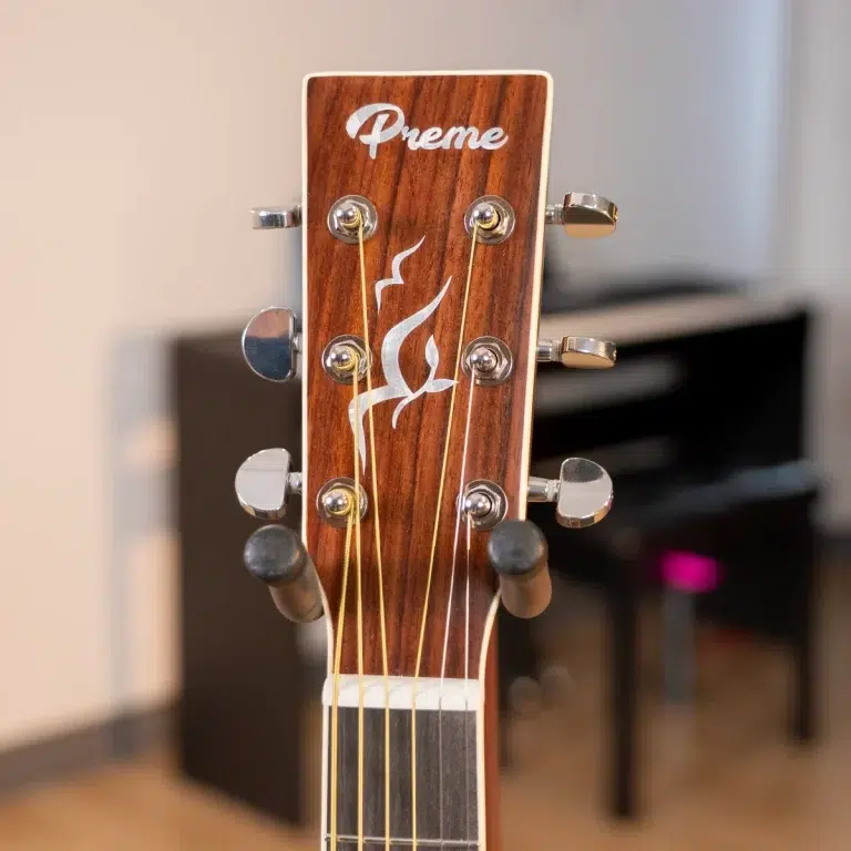 Close-up of a wooden guitar headstock with six tuning pegs and gold strings, a white flame inlay and the brand name 'Preme' on the headstock. ขายราคาพิเศษ