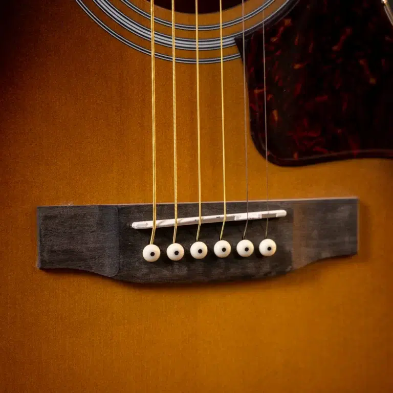 Close-up of an acoustic guitar's bridge with six white bridge pins and a white saddle on a warm-brown body ขายราคาพิเศษ