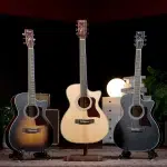 Three acoustic guitars on stands lined up indoors, with a light‑wood center guitar flanked by two darker sunburst guitars; a warm lamp and storage drawers in the background. ลดราคาพิเศษ