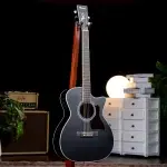 Black acoustic guitar on a stand in a dark studio with a lamp and white drawers in the background, creating a cozy music setup. ขายราคาพิเศษ