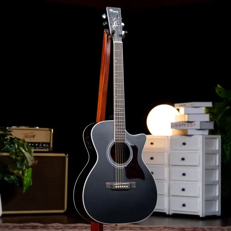 Black acoustic guitar on a stand in a dark studio with a lamp and white drawers in the background, creating a cozy music setup. ขายราคาพิเศษ