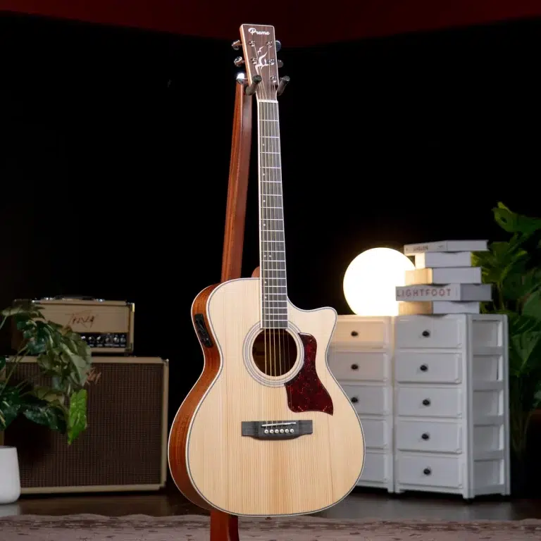 Acoustic cutaway guitar on a stand in a home studio setup with a lamp, white drawers, plants, and an amplifier in the background. ขายราคาพิเศษ