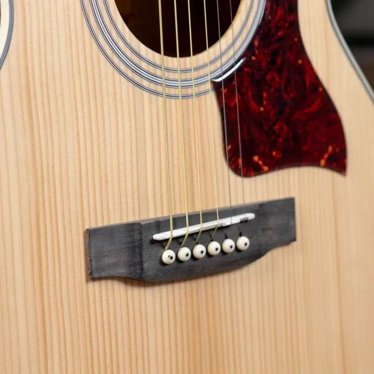 Close-up of an acoustic guitar showing the bridge, six strings, and tortoiseshell pickguard on a light wood body. ขายราคาพิเศษ