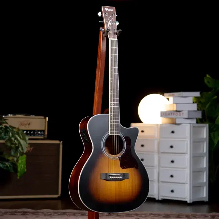 Sunburst acoustic guitar on a stand in a dim room, with a warm light source and white storage drawers in the background. ขายราคาพิเศษ