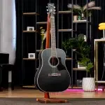 Black acoustic guitar on a wooden stand in a modern showroom with shelves and plants in the background, ขายราคาพิเศษ