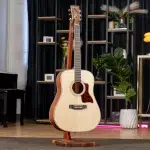 Acoustic guitar on a wooden stand in a modern showroom with shelves, plants, and warm lighting behind it. ขายราคาพิเศษ