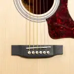 Close-up of an acoustic guitar's bridge with six white bridge pins, saddle, and the light wood body visible near the soundhole and tortoiseshell pickguard. ขายราคาพิเศษ