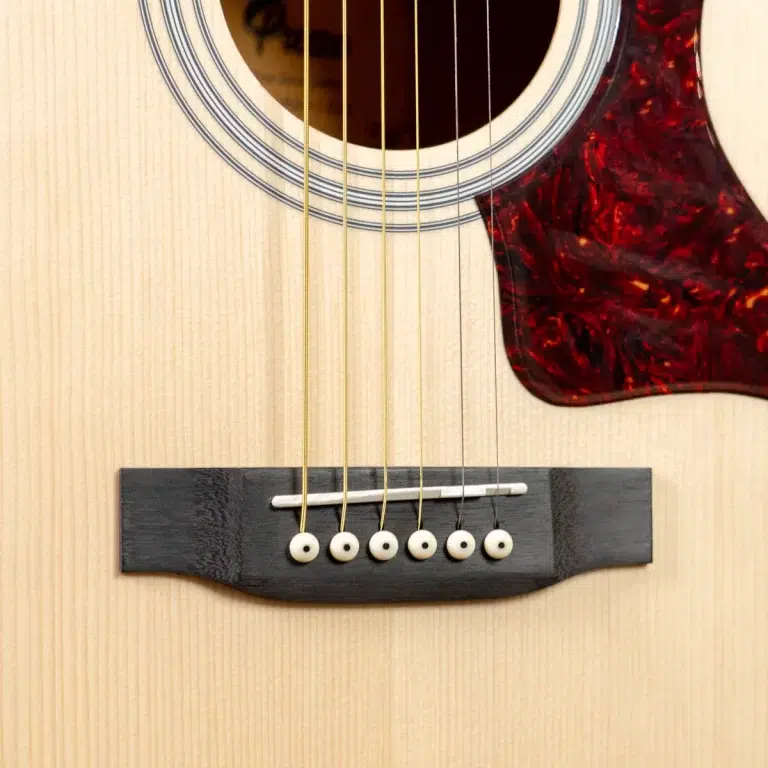 Close-up of an acoustic guitar's bridge with six white bridge pins, saddle, and the light wood body visible near the soundhole and tortoiseshell pickguard. ขายราคาพิเศษ