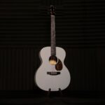 White acoustic guitar on a stand in a dimly lit room with acoustic panels in the background, front view showing the strings and tortoise-shell pickguard area. ลดราคาพิเศษ