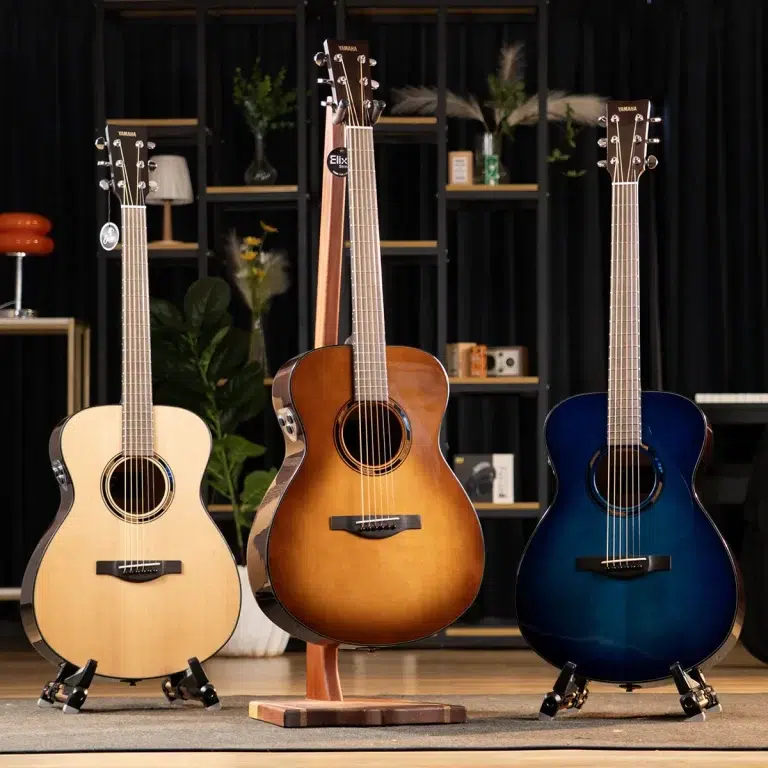 Three acoustic guitars on stands in a music showroom: natural left, sunburst center, and blue right against a shelving backdrop. ขายราคาพิเศษ