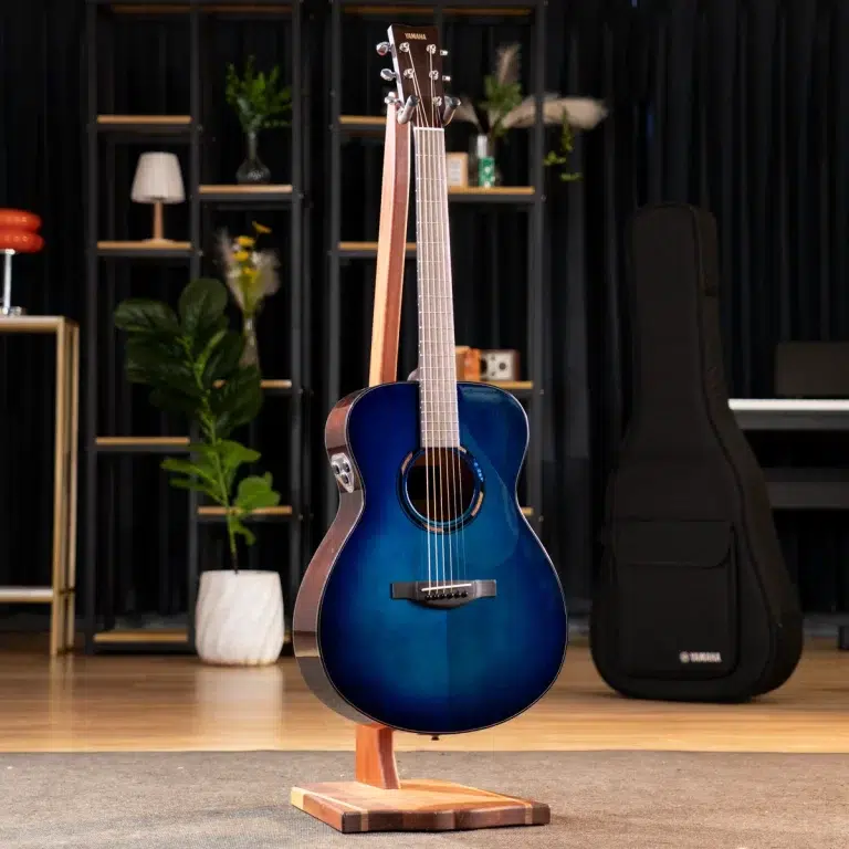 Blue acoustic guitar in a wooden stand, set in a studio with shelves, plants, and a black guitar case in the background, Yamaha logo on the headstock. ขายราคาพิเศษ