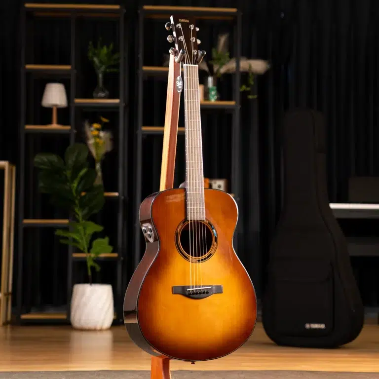 Sunburst acoustic guitar on a stand in a music studio with shelves and a guitar case in the background. ขายราคาพิเศษ
