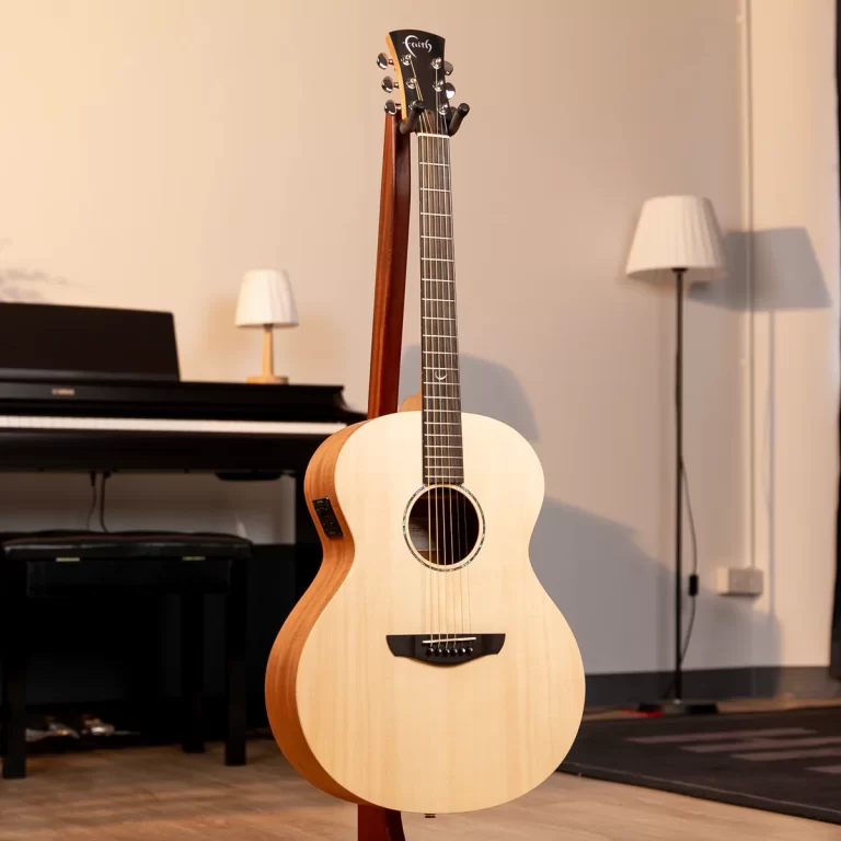 Acoustic guitar on a stand in a cozy room with a keyboard in the background and a floor lamp nearby. ขายราคาพิเศษ