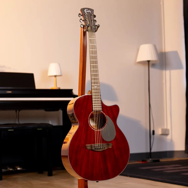 Red acoustic guitar on a stand in a room with a piano and floor lamp behind it, warm indoor lighting ขายราคาพิเศษ