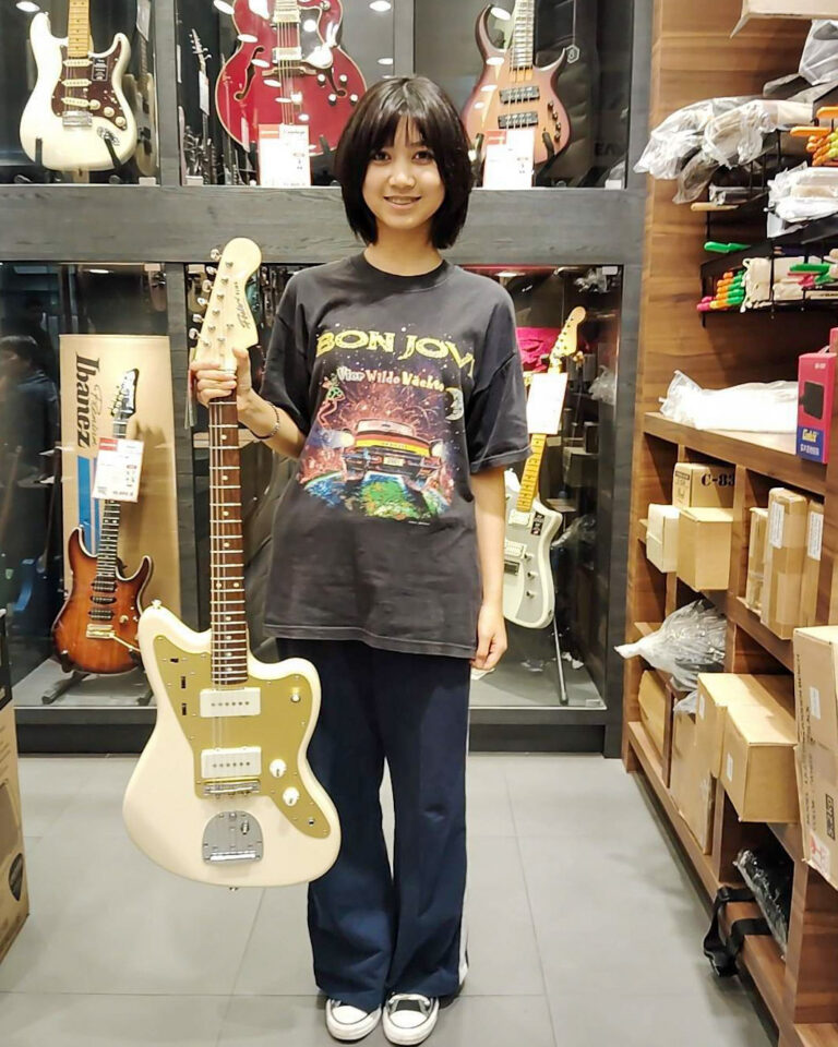 Young woman in a Bon Jovi T-shirt stands in a guitar shop, holding a cream Fender-style electric guitar by its neck.