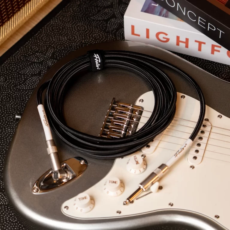 Electric guitar body with a coiled black instrument cable resting on its white pickguard, knobs labeled Tone and Volume nearby, and a book stack in the background. ขายราคาพิเศษ