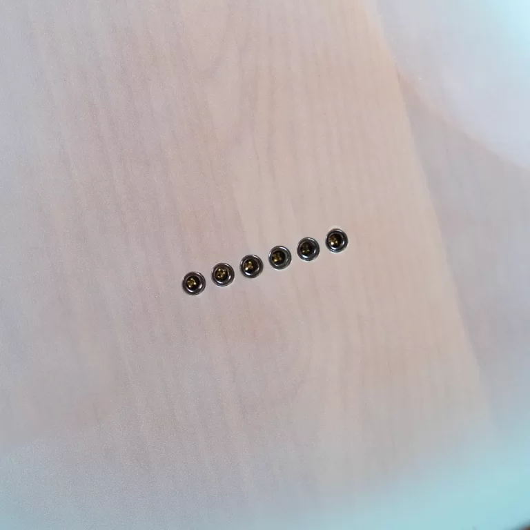 Six small metal grommets or eyelets lined up diagonally on a pale wooden surface. ขายราคาพิเศษ