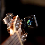 Close-up of a guitar headstock with a Galux AMT-100 digital tuner mounted on the neck, displaying tuning readout in blue and green. ขายราคาพิเศษ