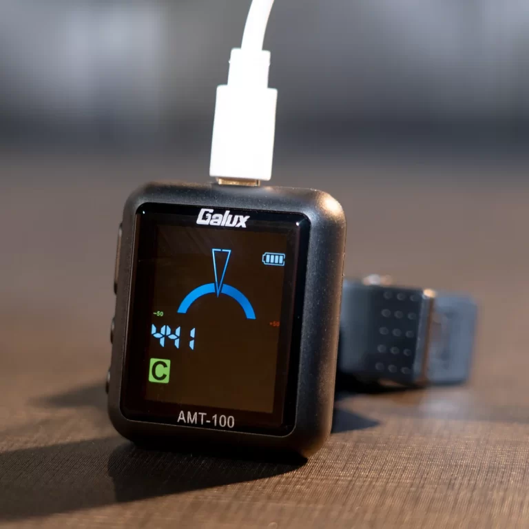 Small black Galux AMT-100 meter with a blue gauge on its screen, plugged into a white USB cable, resting on a brown surface. ขายราคาพิเศษ