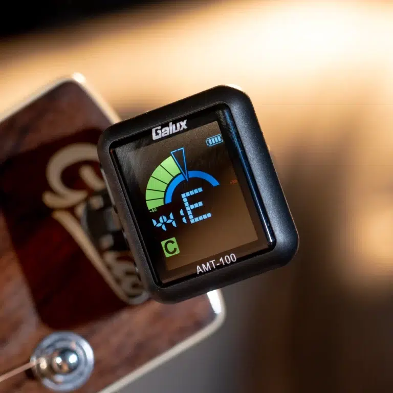 Clip-on guitar tuner mounted on a wooden guitar, showing a blue/green tuning dial and AMT-100 label ขายราคาพิเศษ