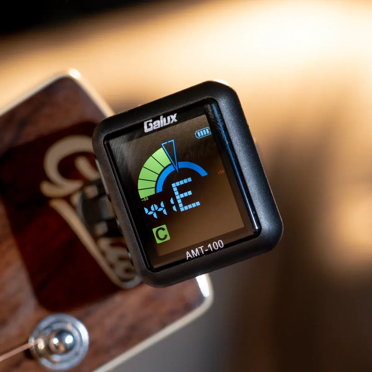 Clip-on guitar tuner mounted on a wooden guitar, showing a blue/green tuning dial and AMT-100 label