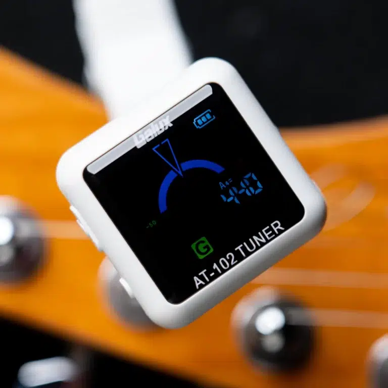 White handheld guitar tuner on a wooden surface, displaying AT-102 TUNER and a blue tuning arc on the screen ขายราคาพิเศษ