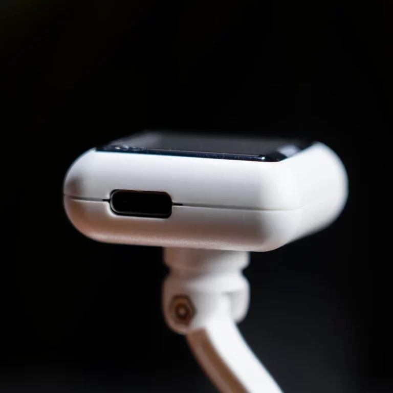 Close-up of a white smartwatch on a stand, showing the side charging port against a dark background. ขายราคาพิเศษ