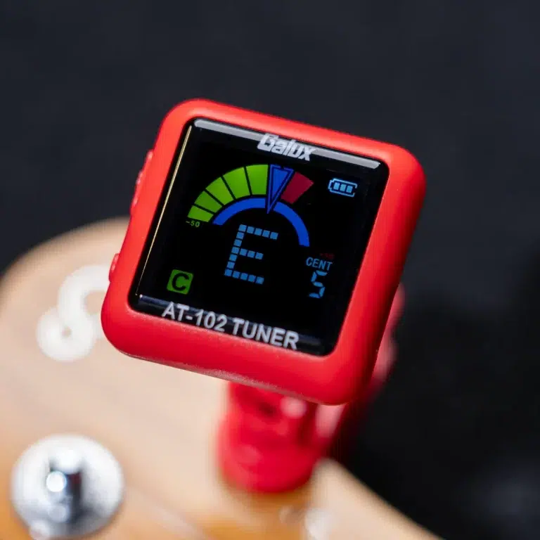Close-up of a red AT-102 tuner with a digital display showing note E and a colorful tuning meter (green arc and blue indicators). ขายราคาพิเศษ
