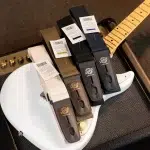 White guitar body with five fabric strap samples in beige, brown, olive, and navy, each with leather ends and tags, resting on a textured amp cabinet. ลดราคาพิเศษ
