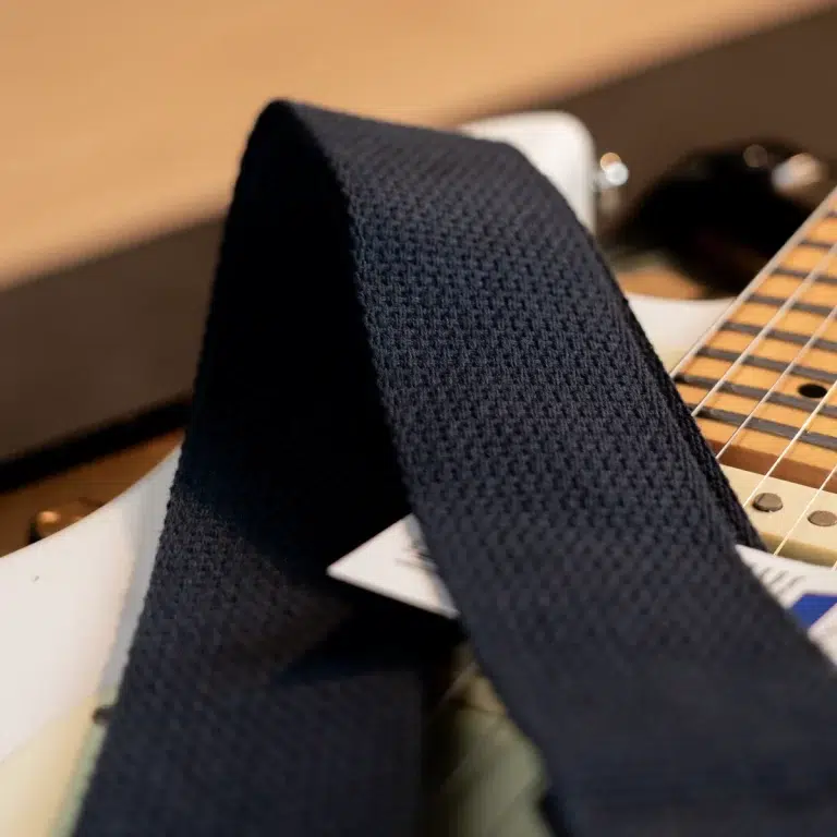 Close-up of a navy woven guitar strap resting over a white electric guitar body and maple fretboard. ขายราคาพิเศษ