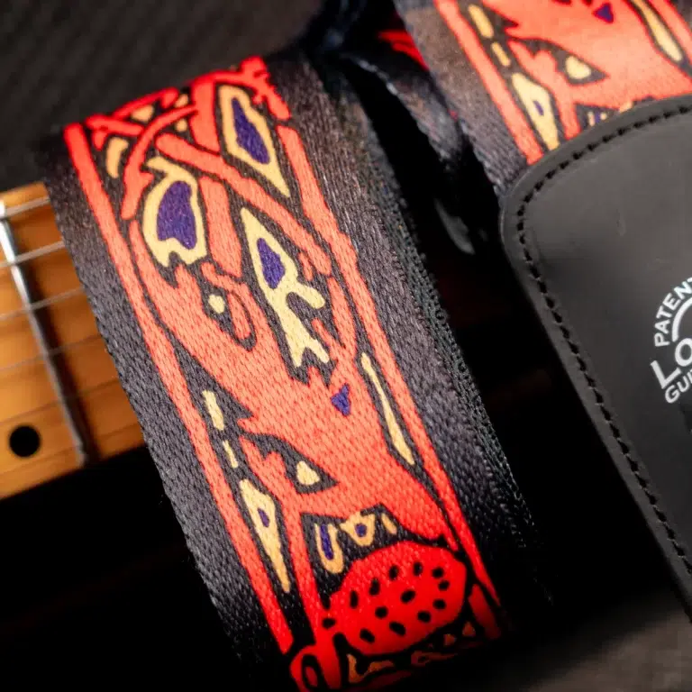 Close-up of a red-orange patterned guitar strap with yellow and purple accents, leather end tab visible beside a guitar neck. ขายราคาพิเศษ