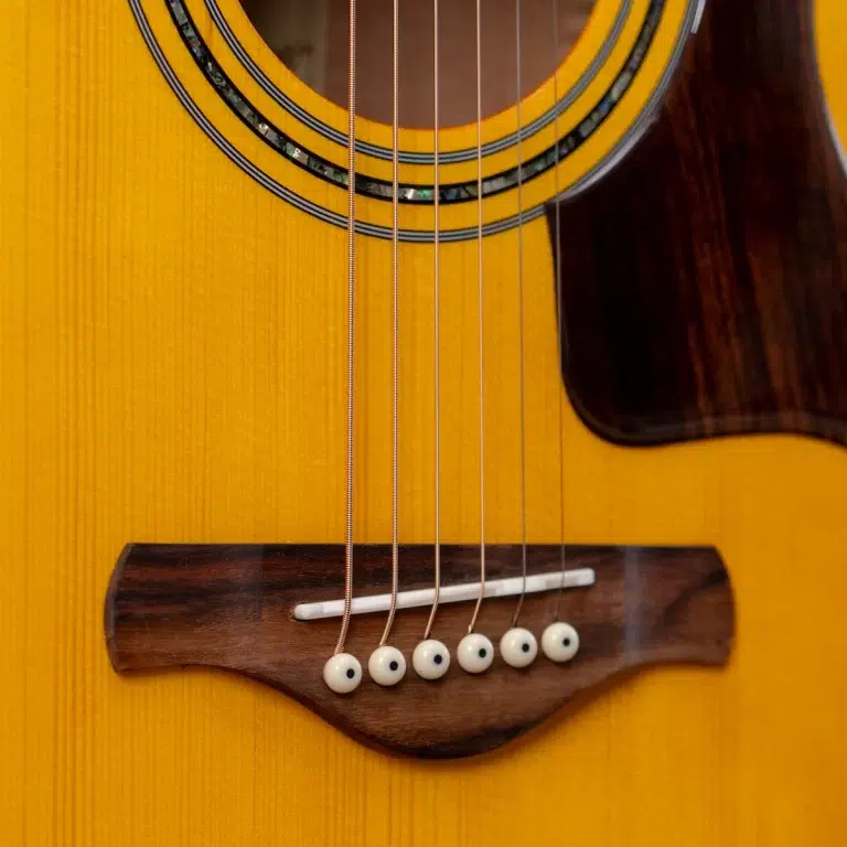 Close-up of an acoustic guitar’s body showing six strings over the dark wooden bridge on a yellow top with a round soundhole rim ขายราคาพิเศษ