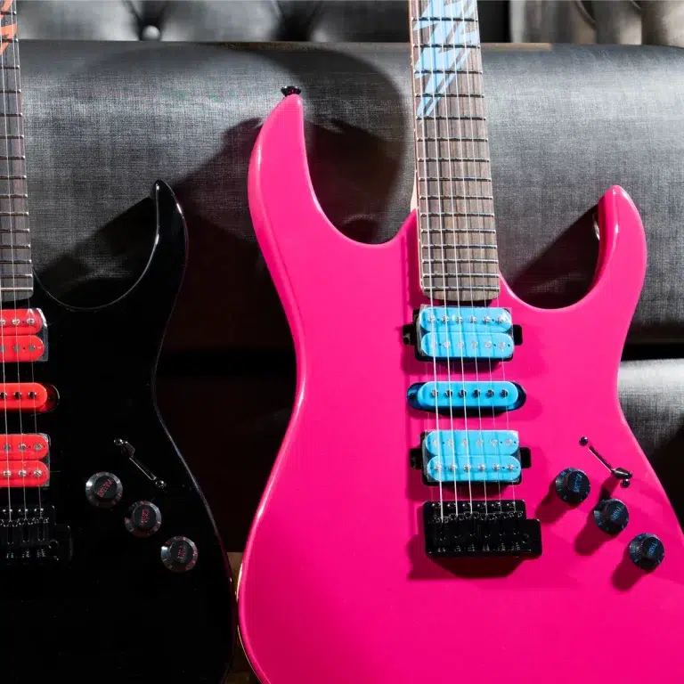 Two electric guitars leaning on a dark sofa: one black with red pickups and knobs, the other bright pink with blue pickups. ขายราคาพิเศษ