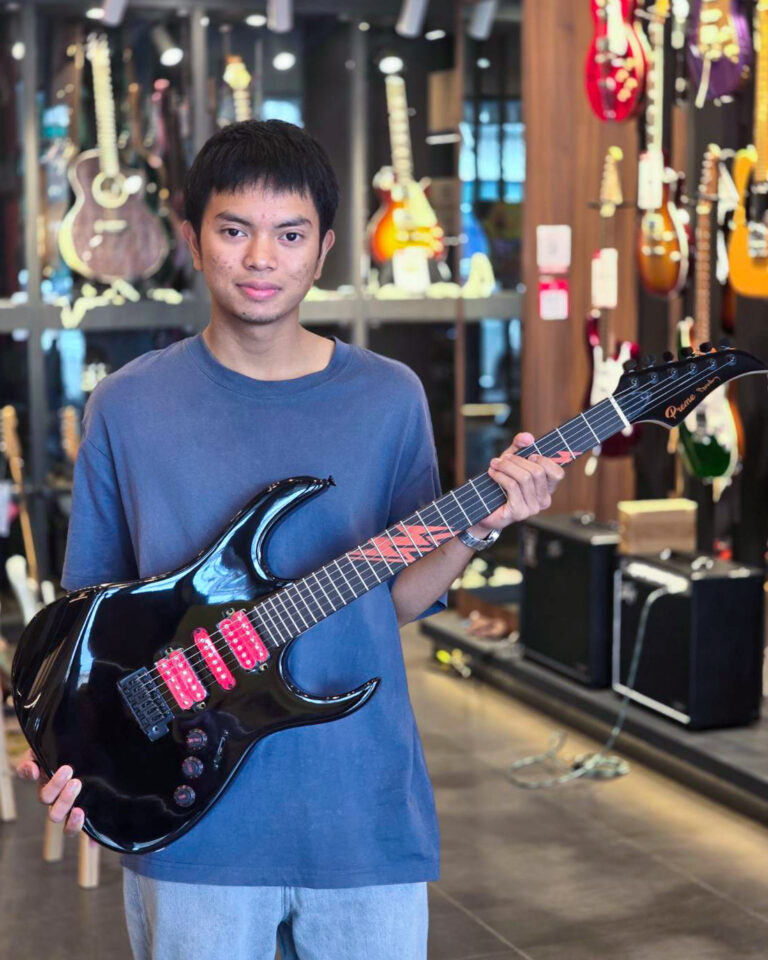 Young man in a blue shirt holding a glossy black electric guitar with pink pickups inside a guitar shop.