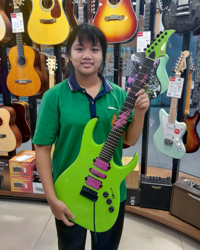 Girl in a green polo shirt holding a bright neon green electric guitar in a music store with guitars on display behind her.