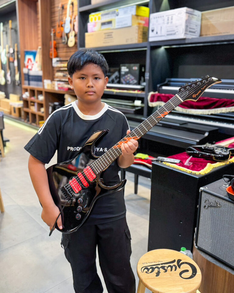 Young boy in a black shirt holding a black electric guitar with red pickups in a music shop. Behind him are shelves of instruments and gear.
