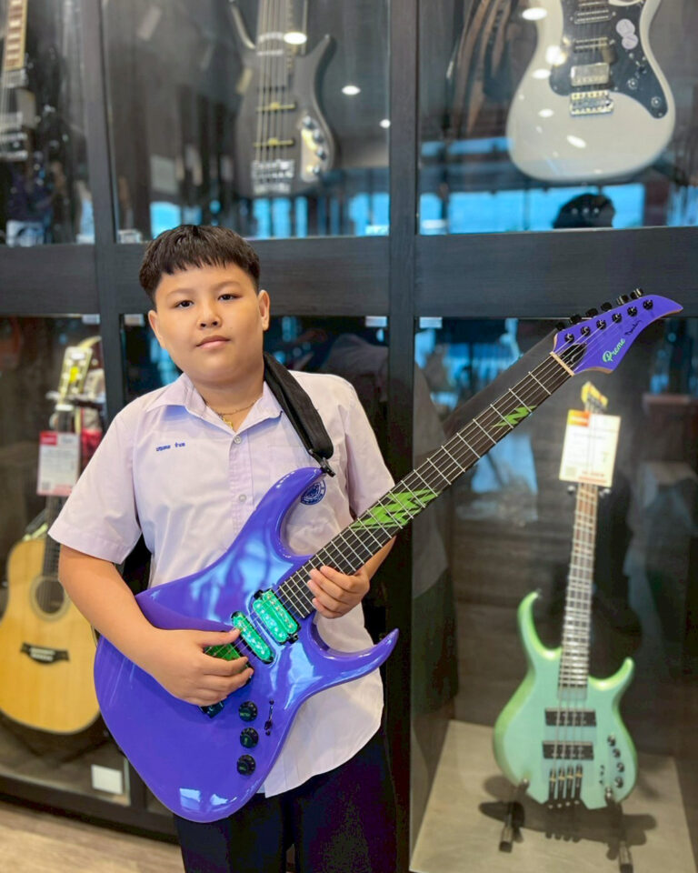 Boy in a white shirt posing with a purple electric guitar in a music store display area with other guitars behind him.