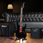 Black electric guitar on a stand in a studio, with a Marshall amplifier and a tufted black sofa in the background. ขายราคาพิเศษ