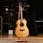 Acoustic guitar on a wooden stand in a studio with shelves, plants, and a piano in the background. ขายราคาพิเศษ