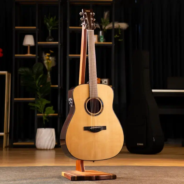 Acoustic guitar on a wooden stand in a studio with shelves, plants, and a piano in the background. ขายราคาพิเศษ