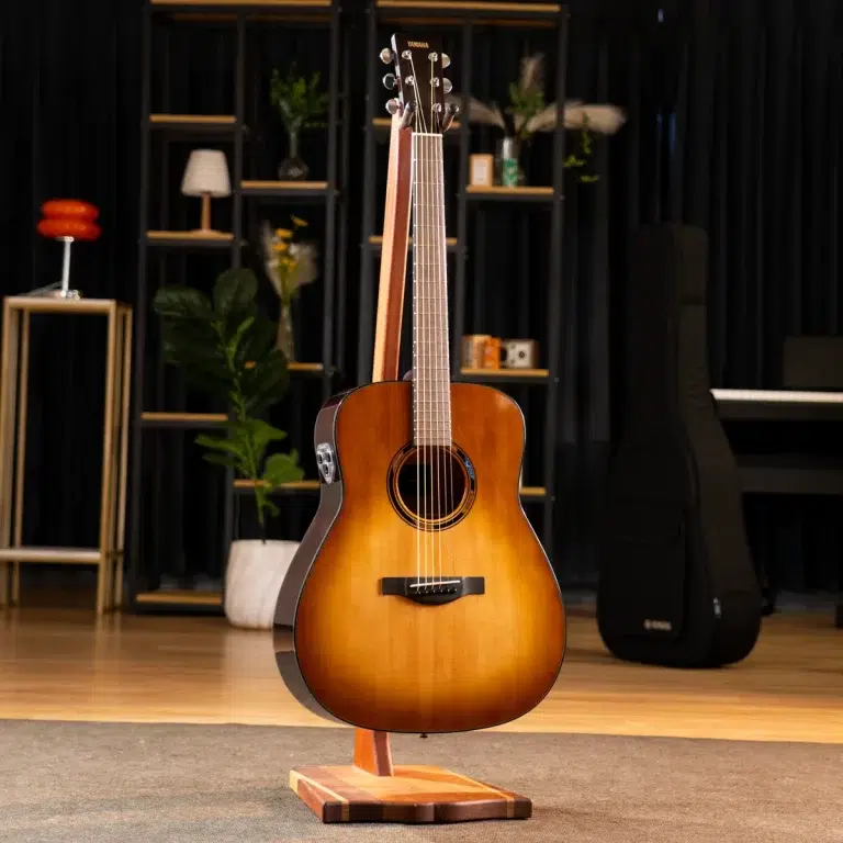 Yamaha acoustic guitar with sunburst finish on a wooden stand in a studio showroom, with shelves and a piano in the background. ขายราคาพิเศษ