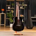 Black acoustic guitar on a wooden stand in a showroom with shelves, plants, and a guitar case in the background. ขายราคาพิเศษ