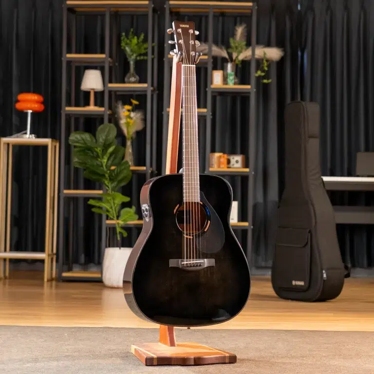 Black acoustic guitar on a wooden stand in a showroom with shelves, plants, and a guitar case in the background. ขายราคาพิเศษ