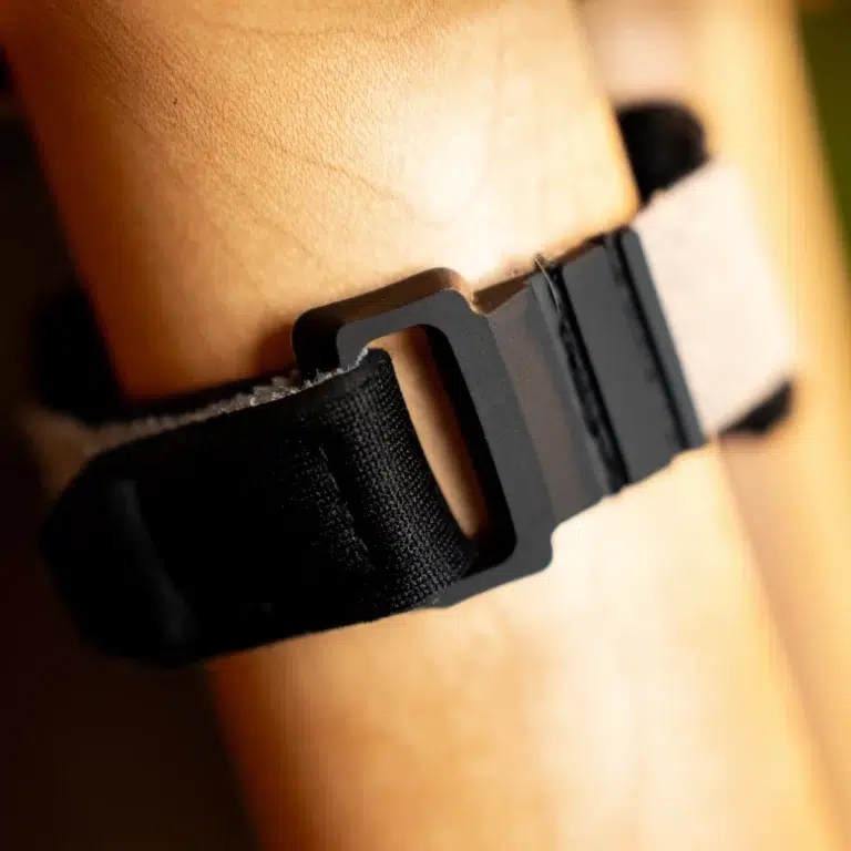 Close-up of a black fabric strap with a gray plastic buckle lying on a wooden surface, showing the adjustable fastening mechanism. ขายราคาพิเศษ