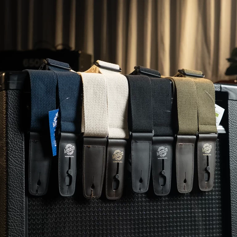 Rows of guitar straps in blue, beige, dark blue, gray, and olive hanging over a black amplifier. Leather ends with buckle closures are visible. ขายราคาพิเศษ