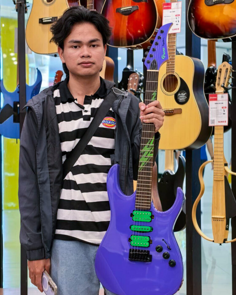 Young man in a striped shirt and jacket holds a purple electric guitar in a guitar shop.