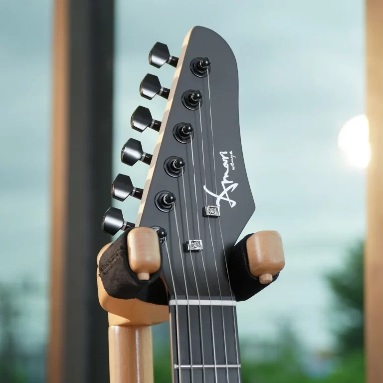 Close-up of a dark gray guitar headstock with six tuning pegs and steel strings, held in a wooden stand, showing a white logo on the headstock. ขายราคาพิเศษ