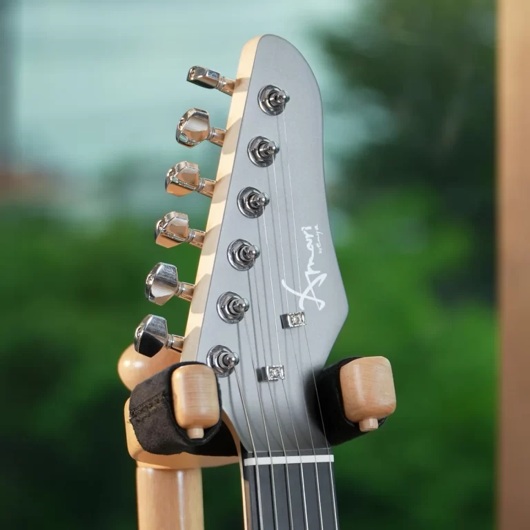 Close-up of a guitar headstock with chrome tuning pegs on a gray finish, mounted on a stand against a blurred green background. ขายราคาพิเศษ