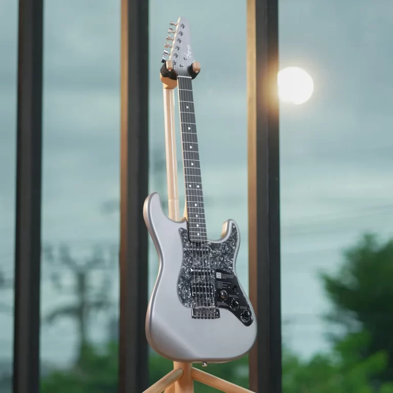 Silver electric guitar on a stand indoors by large windows, sun glare in the background, pearl-like pickguard details visible. ขายราคาพิเศษ