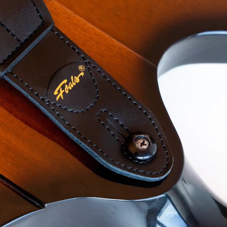 Close-up of a brown leather guitar strap with a gold logo, attached to a strap button on a glossy guitar body. ขายราคาพิเศษ